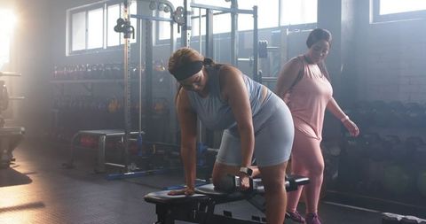 Diverse women performing single-arm dumbbell row with spotter in sunlit gym