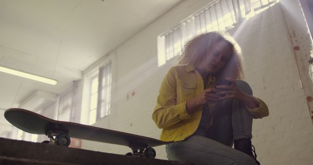 Teen woman with curly hair sitting on indoor urban steps with skateboard beside her, engaging with smartphone under natural light. Great for urban lifestyle projects, tech usage, youth culture promotions, or ads targeting the teenage demographic.