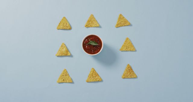 Corn tortilla chips arranged in a geometric pattern around a bowl of salsa dip on a blue background creates an eye-catching and minimalist design. This clean and simple food presentation is perfect for promoting Mexican cuisine, snack foods, restaurant menus, party planning, or any food blog content seeking to showcase clean, organized, and appealing visuals.