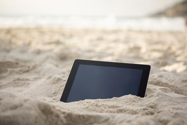 Digital tablet partially buried in sand at beach, representing modern technology in a vacation setting. Ideal for use in travel blogs, technology articles, vacation planning websites, and advertisements promoting digital devices for outdoor use.