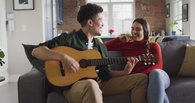 Young couple sits on a comfortable couch in a cozy living room, enjoying music and bonding through an acoustic guitar session. Smiling and engaging in laid-back conversation, conveying warmth and intimacy. Perfect for lifestyle, music, and romance-themed campaigns.