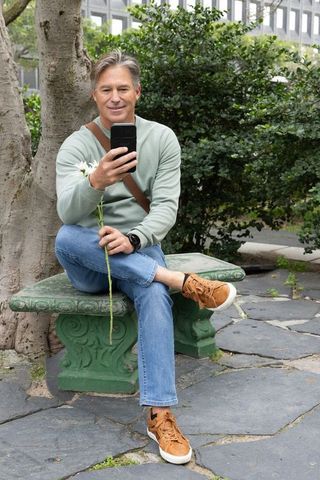 Mature man sitting on green bench holding smartphone and white daisy in urban courtyard