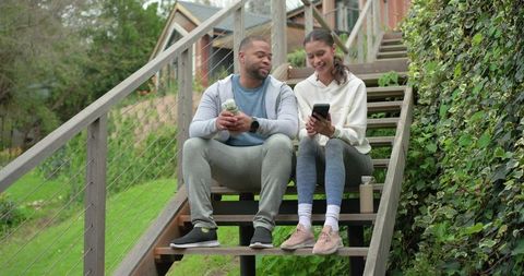 Diverse couple sitting on backyard steps in athleisure sharing smartphone and water bottles