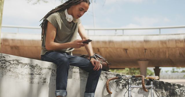 Fashionable biracial man with dreadlocks sitting on wall and using smartphone - Download Free Stock Photos Pikwizard.com