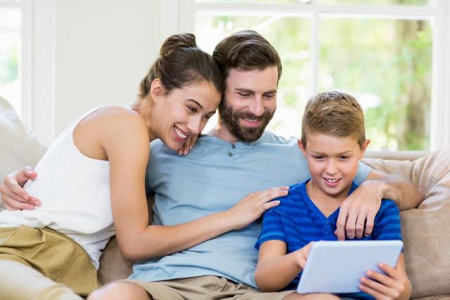 Image shows a happy family using a digital tablet while sitting on a couch in their living room. The father, mother, and son are smiling and looking at the tablet screen together. Perfect for promoting family bonding, technology in the home, and digital learning.