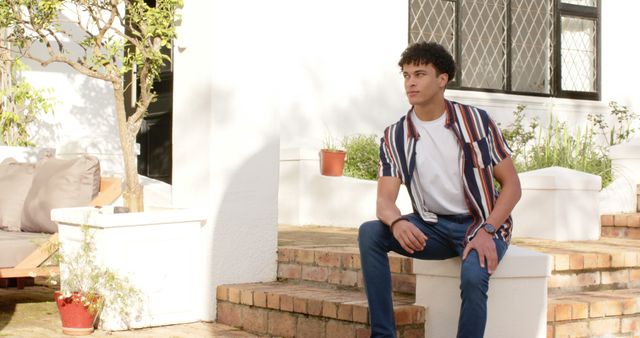 Young man in casual attire sitting outdoors on steps of white home, enjoying sunny day. Great for lifestyle, residential, home ownership, and modern living concepts.