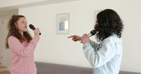 Female friends singing karaoke duet in cozy living room holding microphones and laughing