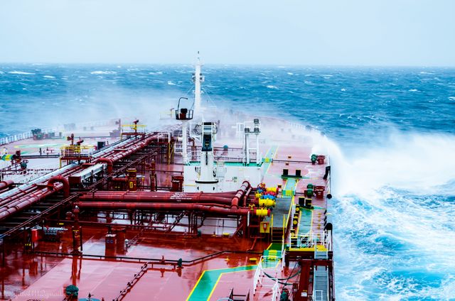 Large cargo ship experiencing rough seas in open ocean with waves crashing on deck. Useful for illustrating maritime conditions, shipping and transportation, and challenges in marine navigation. Ideal for articles or publications dealing with sea freight, marine weather conditions, and nautical themes.