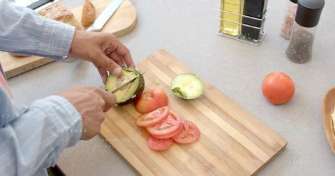 Hands slicing avocado on wooden cutting board with tomato slices for fresh sandwich