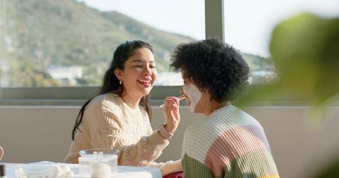 Women applying facial mask together by window, enjoying self care ritual