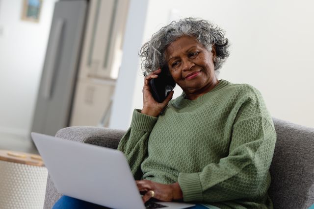Senior African American woman sitting on couch, talking on phone while using laptop. Ideal for illustrating concepts of remote work, senior lifestyle, communication, and inclusivity. Suitable for use in articles about elderly technology use, home office setups, and senior independence.