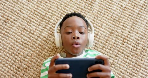 Young african american boy lying on rug wearing headphones playing on smartphone