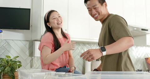 Asian couple laughing while packing clear storage bins in bright modern kitchen