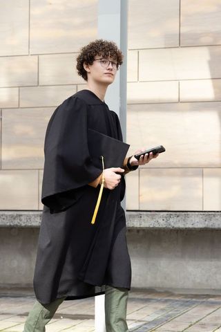 Male graduate walking on campus holding mortarboard with yellow tassel and smartphone