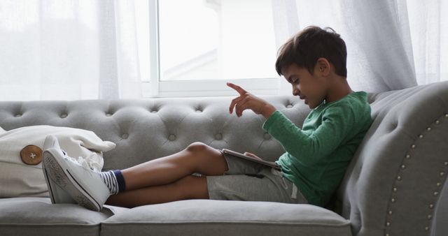 Young boy in casual clothes sitting on gray sofa using tablet. Light coming through window, creating a calm atmosphere. Ideal for themes related to childhood, technology usage among children, home settings, and relaxation. Perfect use cases include parenting blogs, technology-related articles, childhood education materials, and advertisements for casual furniture or children's tech products.