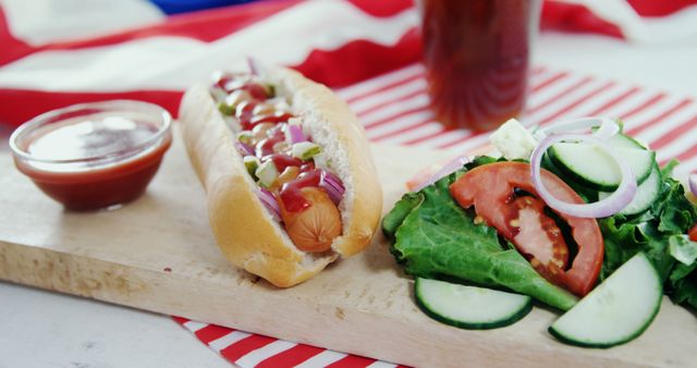 Hot dog with ketchup, mustard, onions, and pickles on bun next to fresh veggie salad with lettuce, tomato, baking paper over table. Can be used for summer cookout, picnic advertisement, food documentaries, casual dining promotions.