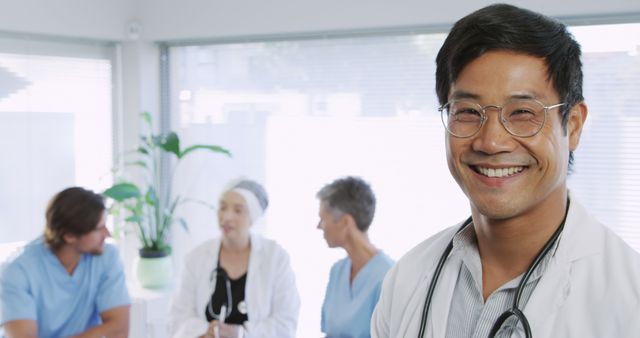 Image shows a smiling Asian male doctor wearing a white coat and stethoscope standing in a hospital with a medical team engaged in a conversation in the background. Ideal for promoting healthcare services, showcasing hospital staff, or illustrating teamwork in a medical context on websites and marketing materials.