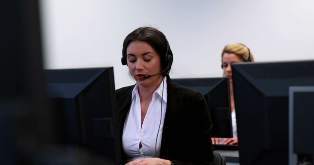Caucasian women in professional attire are working in a call center environment, with one focused on her computer screen wearing a headset. Their engagement suggests a busy customer service department, highlighting the importance of communication in business operations.