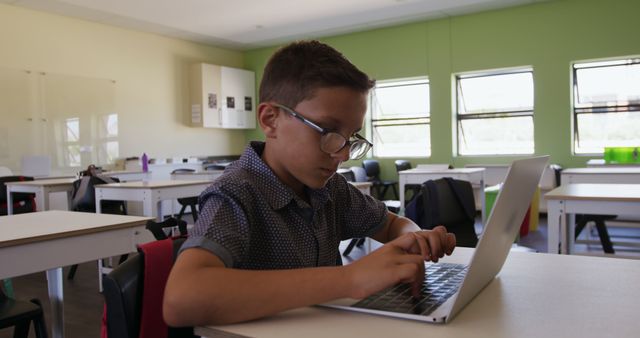 Young boy focused on laptop in brightly-lit classroom with green walls. Ideal for educational materials, technology in schools, youth learning, academic enviroments, and back-to-school promotions.