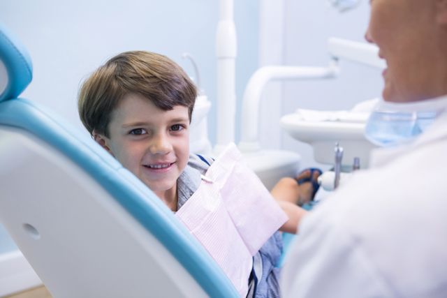 Young boy sitting on dentist chair, smiling at camera. Ideal for use in articles about pediatric dentistry, dental care for children, healthcare services, and promoting dental hygiene. Can be used in brochures, websites, and advertisements for dental clinics and healthcare providers.