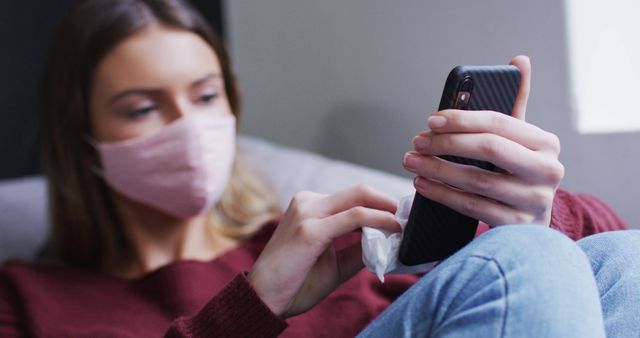 Image shows woman wearing a face mask while using a smartphone and cleaning its screen with a cloth. The importance of hygiene and virus prevention is emphasized. Useful for health-related articles, pandemic awareness campaigns, and technology usage during health crises.