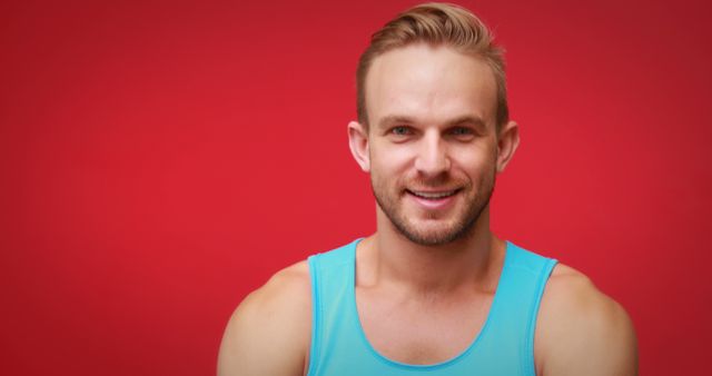 A smiling young Caucasian man wearing a blue tank top stands against a vibrant red background, with copy space. His cheerful expression and casual attire suggest a relaxed and positive atmosphere.