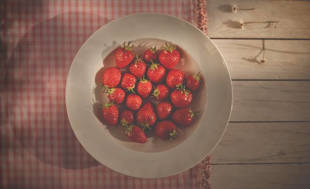 Bright red, fresh strawberries filling white bowl on rustic wooden table with gingham cloth. Perfect for promoting healthy eating, organic food, summer desserts, food blogs, kitchen decor, and cooking inspiration.