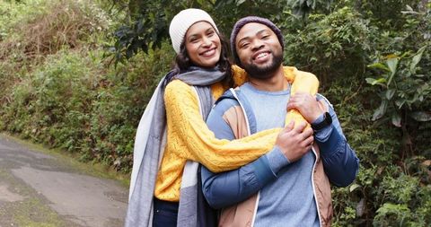 Smiling couple embracing on leafy trail wearing knit beanies and cozy autumn layers