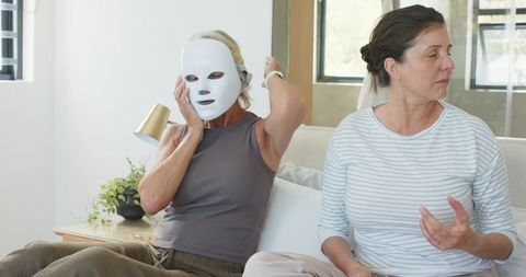 Diverse women wearing led facial masks relaxing and talking on sofa