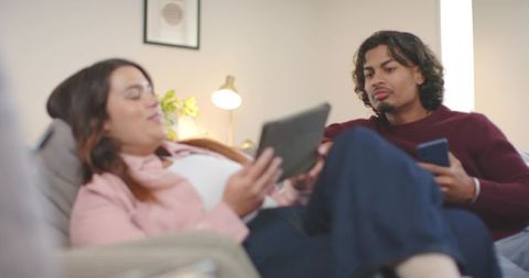 Young couple lounging on sofa using tablet and phone, relaxing in cozy living room