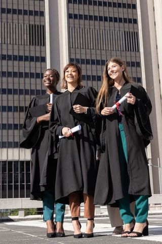 Sunlit multicultural female graduates posing in urban academic gowns holding diplomas