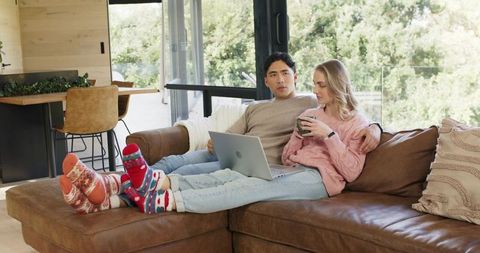 Young couple relaxing on leather couch using laptop and sipping coffee in cozy living room