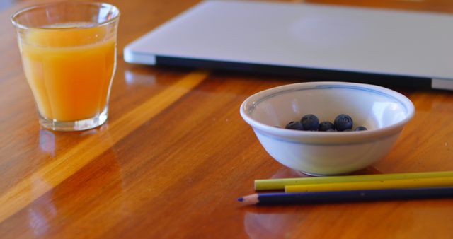 Image showcasing a healthy workspace with a glass of orange juice, bowl of blueberries, and a laptop on a wooden desk. Also, there are pencils laid out on the desk, indicating an organized and productive environment. Ideal for articles or advertisements on remote working, healthy office habits, work-life balance tips, productivity, and nutritious snacks.