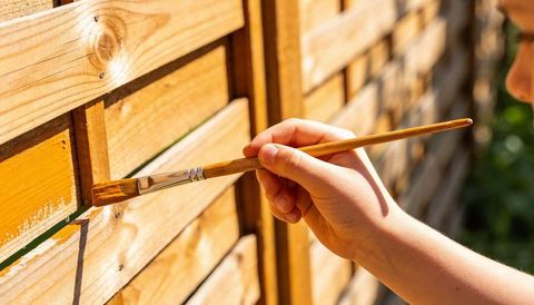 Staining wooden slatted fence with brush under warm sunlight, close-up outdoor DIY project