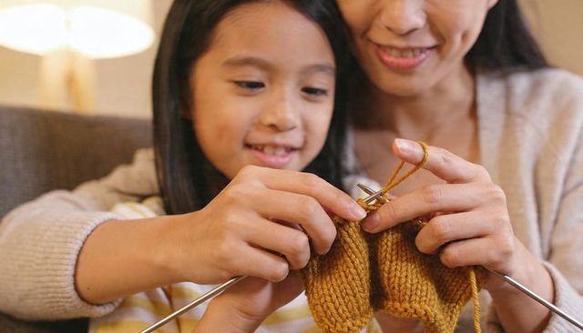 Mother and daughter knitting together on sofa using metal needles and mustard yarn