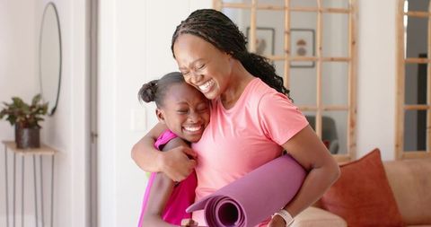 African american mother and teen daughter hugging with yoga mat in sunlit living room
