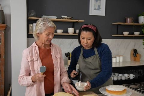 Mature women decorating layer cake together in cozy modern kitchen, baking and teamwork