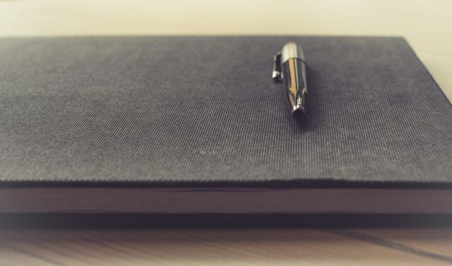 Journal placed on desk with metal pen on top, ideal for themes related to writing, planning, organization, and professional office settings. Suitable for use in blog posts about productivity, motivational articles, or designs for business stationery.