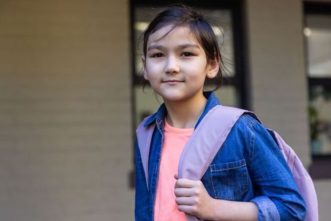 Confident asian schoolgirl standing at school entrance wearing denim jacket and backpack