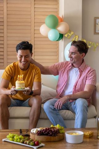 Male couple celebrating birthday at home holding cake on sofa with balloons and snacks