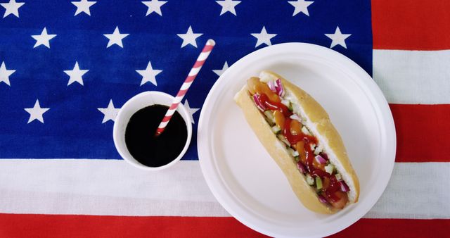 White plate holding hot dog with toppings such as ketchup, mustard, pickles, and onions, soda in cup, all set on top of US flag-themed surface. Great for articles or advertisements about American culture, 4th of July celebrations, traditional American foods, and festive events.