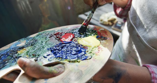 This image shows an artist mixing colors on a wooden paint palette using a brush. Bright colors and artistic materials are prominently featured, making it ideal for art-related content, tutorials on painting techniques, or emphasizing the creative process in marketing visuals.