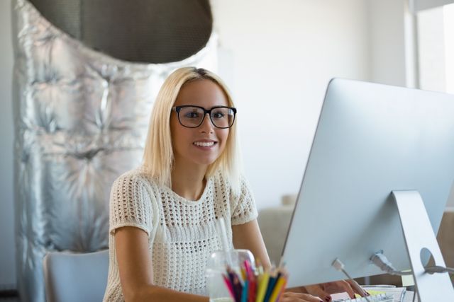 Young woman with blonde hair and glasses working on a computer in a modern, creative office. She is smiling and appears to be enjoying her work. Ideal for use in articles or advertisements related to modern work environments, productivity, technology in the workplace, or professional women in business.