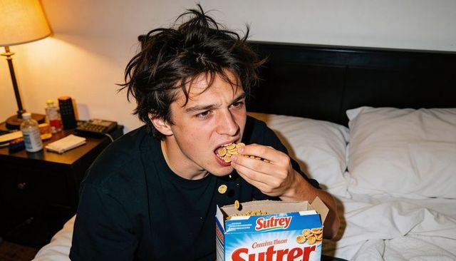 Young man eating cereal on bed holding sutrey box lamp casting warm light late-night snack