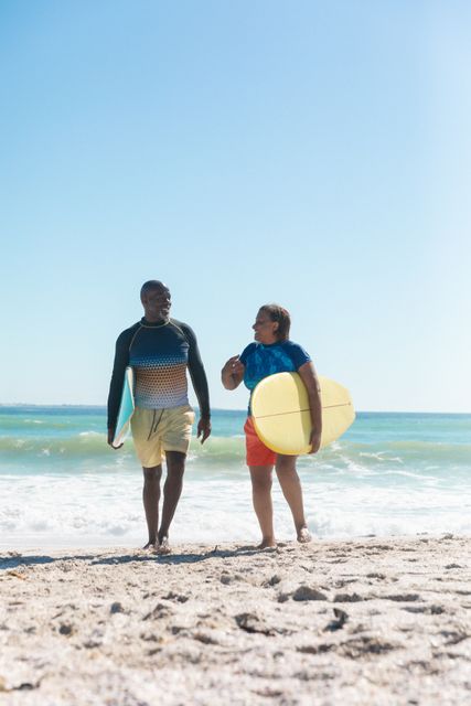 African american senior couple with surfboard walking at beach against blue sky with copy space. unaltered, togetherness, active lifestyle, aquatic sport and holiday concept.