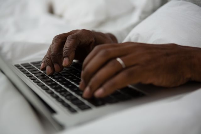 Hands typing on a laptop while sitting on a bed at home. Ideal for illustrating remote work, home office setups, technology use, and casual productivity. Useful for articles or advertisements related to working from home, technology, and comfortable work environments.