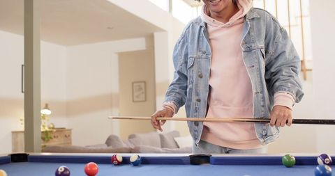 Young man playing pool in modern living room wearing denim jacket and pink hoodie