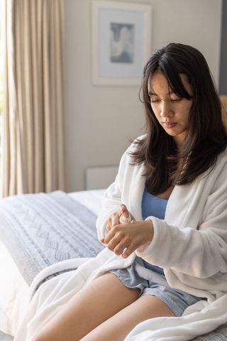 Asian woman applying hand lotion in robe on bed during morning self-care routine