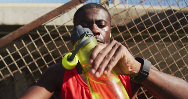 Tired african american man drinking from water bottle, taking break in exercise outdoors. fitness, healthy and active lifestyle concept.