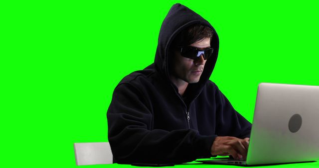 A male Caucasian hacker in a black hood and sunglasses using a laptop, green screen in the background. Computer programming and cyber security. 